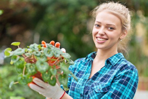 A young woman with light blonde hair tied back, wearing a blue and green checkered shirt and white gardening gloves, is standing outdoors in a garden. She is smiling at the camera while holding a small potted plant with green leaves and some reddish-brown stems. The background features blurred lush greenery, including trees and shrubs, indicating a well-maintained garden space in Kentish Town. The scene suggests active gardening or plant care, with natural daylight highlighting the vibrant colors of the foliage and the woman's cheerful expression, reflecting its relevance to gardening and outdoor landscaping services offered by Landscapers Kentish Town.