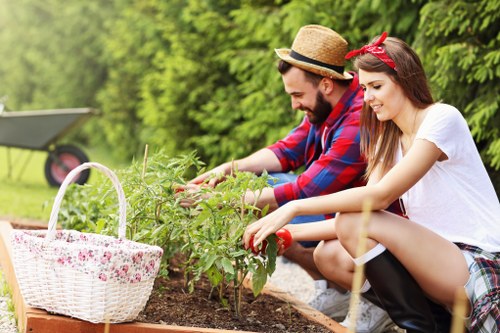 In a well-maintained garden in Kentish Town, two individuals are engaging in outdoor gardening activities; a man wearing a straw hat, plaid shirt, and the woman with long dark hair, a red headband, and casual white t-shirt, are both smiling and carefully tending to a lush green plant in a raised garden bed. The garden bed is filled with healthy, leafy plants and is situated on a neatly prepared soil surface. To the left, a white wicker basket with a floral pattern sits on the edge of the garden bed, possibly holding tools or harvested produce. The background reveals a vibrant environment with tall trees and bushes providing natural shade, along with a paved pathway or patio area. The scene is illuminated by natural daylight, indicating clear, pleasant weather suitable for outdoor gardening. The setting reflects a peaceful and productive outdoor space, highlighting typical features of a residential garden that Landscapers Kentish Town might service, including lawn areas, plant beds, and outdoor leisure zones, reinforcing the contextual relevance to local gardening and landscaping expertise.