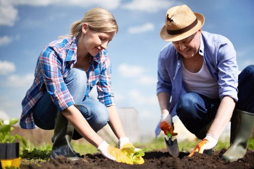 The image depicts a young woman and an older woman working together in a garden, planting or tending to a bed of dark, rich soil. The younger woman, with blonde hair tied back, is wearing a blue plaid shirt and jeans, while the older woman, wearing a wide-brimmed straw hat, a light blue shirt, and dark trousers, is assisting with gardening tasks. Both are using small gardening tools and are dressed in gloves suitable for outdoor soil work. The garden appears well-maintained, with a neatly prepared planting area, and is set against a bright, sunny sky with some scattered clouds. In the background, there are hints of green foliage and possibly other garden features, suggesting a landscaped outdoor space typical of residential or community gardens in Kentish Town or nearby areas. The scene emphasizes active outdoor gardening, natural lighting, and the collaborative effort involved in garden maintenance as part of professional gardening services provided by Landscapers Kentish Town.