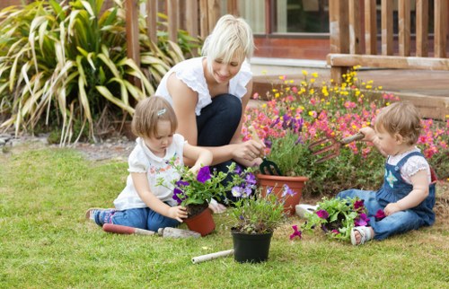A woman and two young children are engaged in gardening in a well-maintained backyard garden, featuring a lush green lawn and a variety of blooming flower plants. The woman, with short blonde hair and casual attire, is sitting on the grass and assisting one child with arranging purple and pink flowers in a small terracotta pot. The children, both with light brown hair and wearing patterned tops and jeans, are actively involved in planting and watering, with additional flower pots placed nearby on the grass. Behind them, there is a dense border of ornamental grasses with long,_striated leaves, providing a natural backdrop and privacy. To the right, a flower bed with bright pink and yellow blossoms lines the base of a wooden patio deck, which has a ramp leading to a house with large windows. This outdoor scene showcases a carefully landscaped yard ideal for family gardening activities, summer weather, and outdoor leisure, embodying professional gardening and landscaping services offered by Landscapers Kentish Town, with natural tones of green, vibrant floral colours, and earthy surfaces contributing to a welcoming and well-kept outdoor environment in London postcodes.