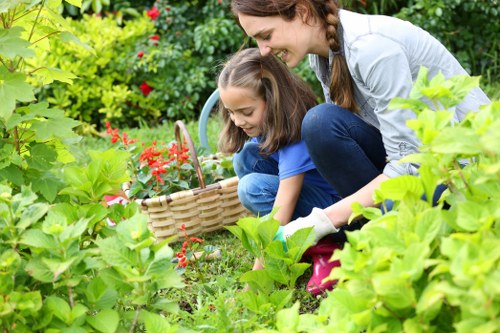 A woman and a young girl are engaging in gardening activities in a well-maintained outdoor space, with lush green foliage and vibrant flowering plants surrounding them. The woman, dressed casually with long brown hair tied back, is crouched down beside the girl, who is wearing a blue T-shirt and jeans, helping her to pick flowers. They are focusing on a flower bed bordered by low greenery, featuring a variety of plants with green leaves and bright red blossoms. In front of them, a woven basket is placed on the grass, possibly for collecting flowers or weeds. The lawn area in the foreground appears dense and healthy, with evenly cut grass. Towards the background, there are well-trimmed hedges and taller trees providing shade and privacy, under bright, natural daylight indicating fair weather. The scene exemplifies outdoor garden care and family-friendly gardening activities, reflecting professional landscaping and gardening services offered by Landscapers Kentish Town in the area around Kentish Town, with a focus on plant care, garden maintenance, and creating inviting outdoor environments.