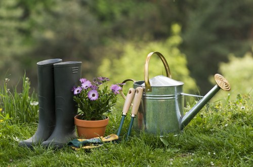 In a well-maintained garden, there are a pair of black rubber wellington boots standing upright on a lush green lawn in the foreground. Next to them, a small terracotta pot contains vibrant purple flowers, adding colour to the outdoor space. A set of garden tools, including a pair of pruning scissors and a small hand rake, are neatly arranged on the grass nearby. A classic metal watering can with a wooden handle and a long spout sits prominently to the right, suggesting active watering or general garden maintenance. The background features softly blurred trees and natural foliage, indicating a peaceful outdoor environment typical of a landscaped garden in Kentish Town. The scene is lit by natural daylight, with no signs of rain or harsh weather, emphasizing a calm, outdoor gardening setting suitable for landscaping and lawn care services. The overall arrangement highlights garden elements like the well-kept grass, decorative flower pot, and essential tools, reflecting a typical gardening activity or preparation in a pleasant, residential outdoor area. This detailed description supports accessibility and local gardening service content for Landscapers Kentish Town, located in postcode NW5.