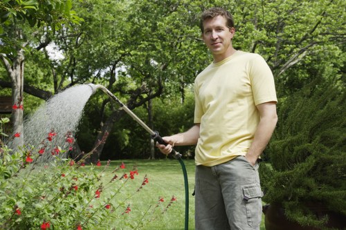 A man standing in a lush garden in Kentish Town, watering a vibrant flower bed with a garden hose on a sunny day. The garden features neatly maintained green grass, a variety of bushes, and tall trees providing natural shade. The flower bed contains red and pink flowers, bordered by soil, with some plants bordering paved pathways. Behind the man, there are well-trimmed hedges and a mixture of deciduous trees with fresh green leaves, indicating a spring or summer season. The scene is well-lit with natural sunlight, emphasizing the healthy foliage and vibrant plant colours. This outdoor space appears to be a well-kept residential garden, managed by local gardening services such as Landscapers Kentish Town, who specialise in lawn care, planting, and garden maintenance to enhance curb appeal and outdoor functionality.