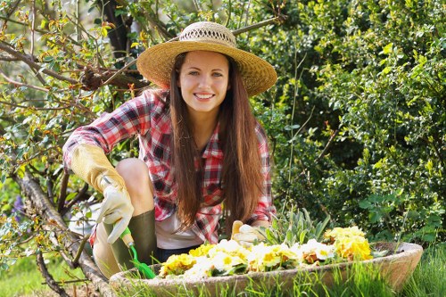 A young woman with long brown hair wearing a straw sunhat, gardening gloves, and a red plaid shirt is kneeling on the grass in a garden setting. She is tending to a large, curved flower bed that contains a variety of blooming flowers in shades of yellow, white, and purple. Behind her, there is a lush backdrop of green foliage, including bushes and small trees, with sunlight filtering through the leaves, indicating a bright, clear day. The grass in the garden is well-maintained and dense, with a smooth, even surface, and the flower bed appears to be bordered by soil or mulch to contain the plants. The environment suggests a well-kept residential garden, suitable for professional gardening and landscaping services by Landscapers Kentish Town, as the woman appears engaged in planting or weeding activities aimed at enhancing outdoor aesthetic appeal.