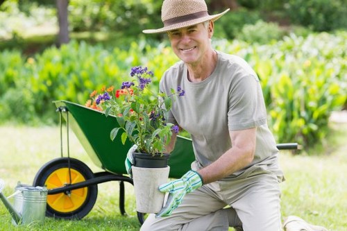 A middle-aged man wearing a wide-brimmed straw hat, grey t-shirt, and gardening gloves is kneeling on the grass in a well-maintained garden in Kentish Town. He is smiling while holding a potted flowering plant with purple and red blooms, preparing to place it in the garden's flower bed or container arrangement. Behind him, a bright yellow and black wheelbarrow filled with garden tools and supplies is positioned on a clean, lush lawn with evenly cut grass. The background features a mix of green foliage, including a hedge or shrubbery, and mature trees that provide dappled shade. The garden displays a tidy, landscaped outdoor area with a natural, inviting atmosphere suitable for outdoor maintenance or landscaping work. The scene captures a peaceful, sunny day, emphasizing the importance of garden care and landscaping services offered by Landscapers Kentish Town, with subtle reference to local outdoor living and gardening enhancements in the area.