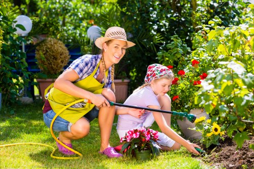 A woman and a girl are gardening together in a lush outdoor space, with the woman crouching and smiling while wearing a straw hat, blue checkered shirt, yellow apron, and pink gardening shoes, holding a yellow garden hose. The girl is kneeling on the grass, wearing a floral headscarf, light-colored shirt, and pink shoes, actively tending to a flower bed with vibrant red, yellow, and pink flowers. The garden features a neatly maintained lawn, various green shrubs and trees in the background, and decorative garden lights hanging above, creating a cheerful and well-tended outdoor environment typical of a landscaped backyard. The scene is set in bright daylight, suggesting warm weather, emphasizing the natural tones of the plants and the well-organized outdoor space associated with gardening services in Kentish Town.
